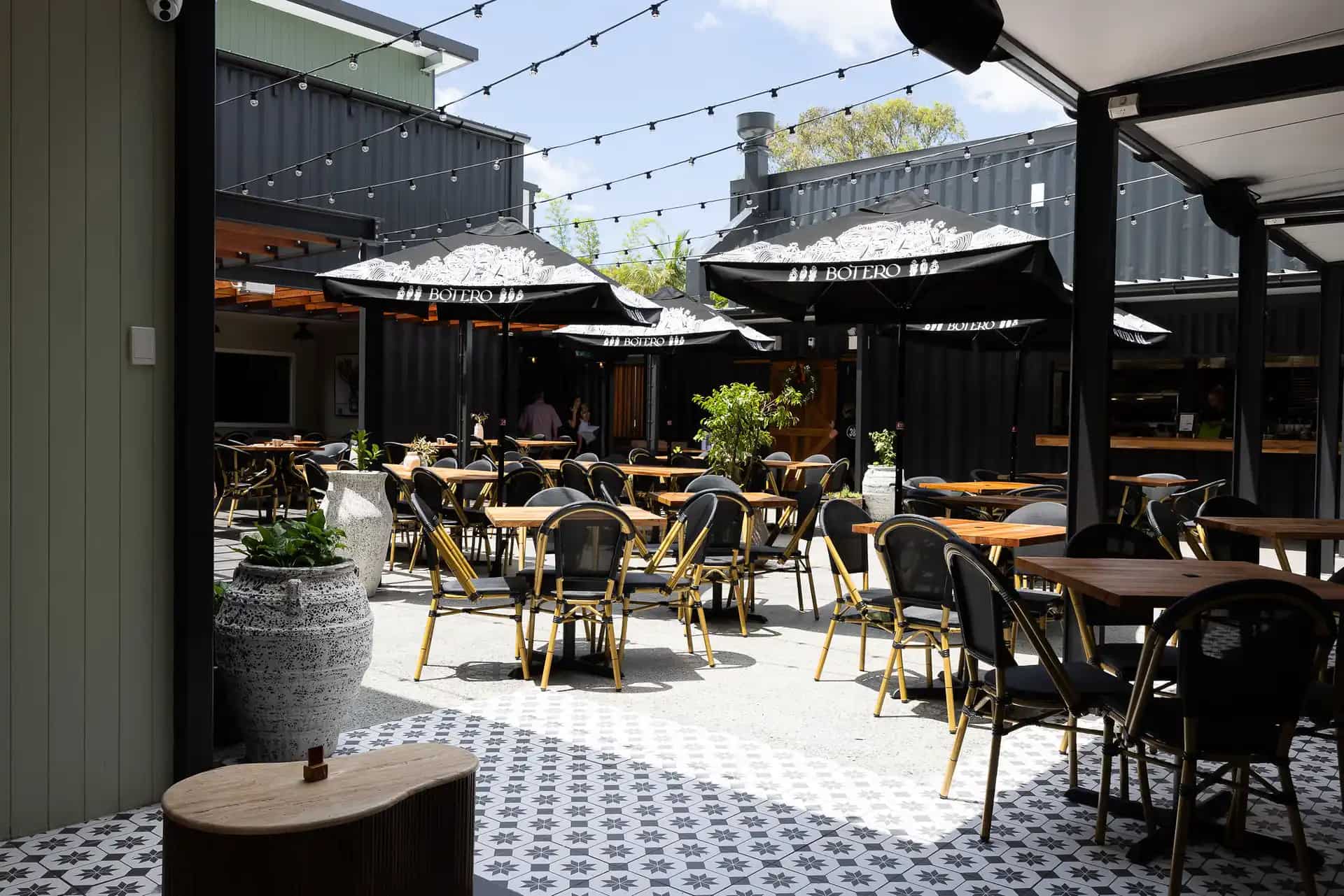 The 383's open-air courtyard: timber tables and rattan bistro chairs under black market umbrellas and festoon lights, ringed by charcoal shipping containers on patterned tiles.