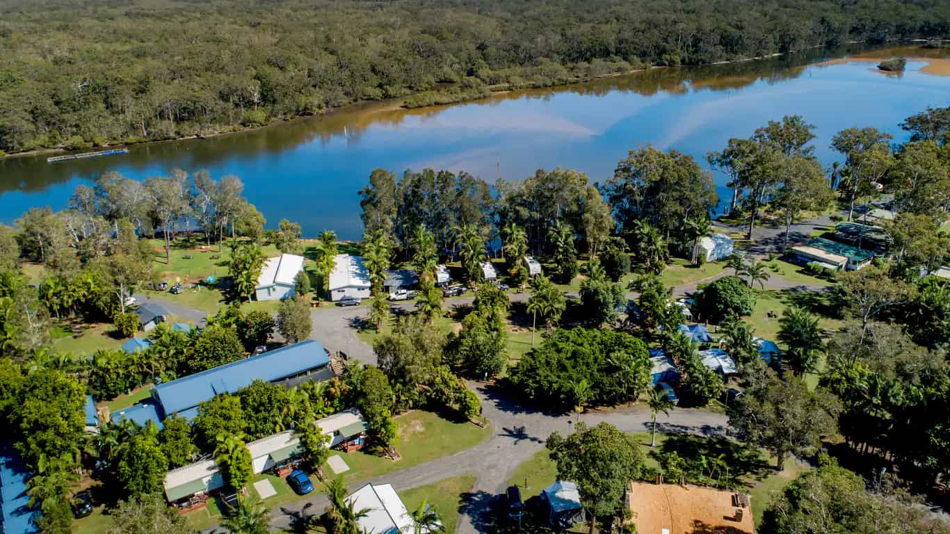 Aerial view of the resort's cabins and caravan sites among palms on the bank of the Wooli Wooli River, with bushland and hills beyond.