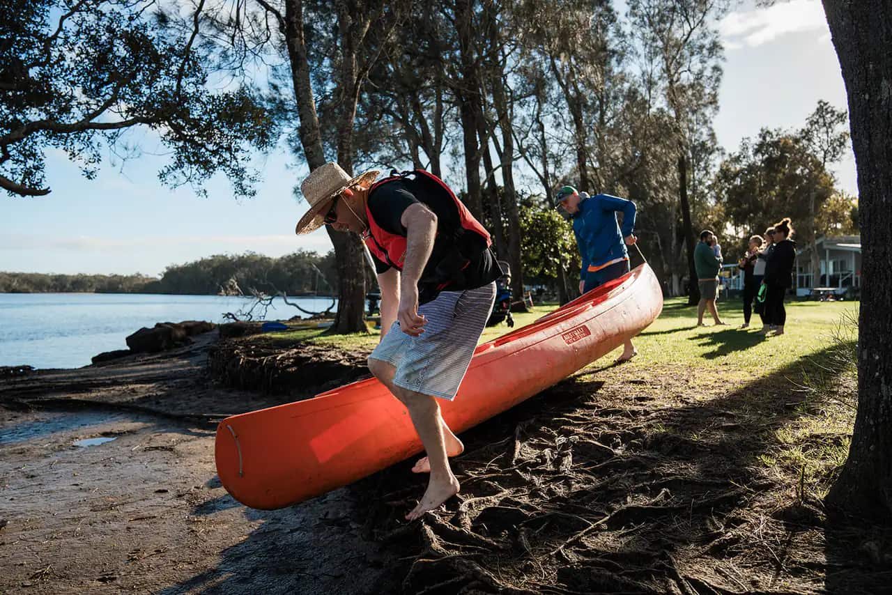 A barefoot crew member in a straw hat and life vest carrying an orange canoe down the tree-shaded riverbank, with others gathered by the water behind.