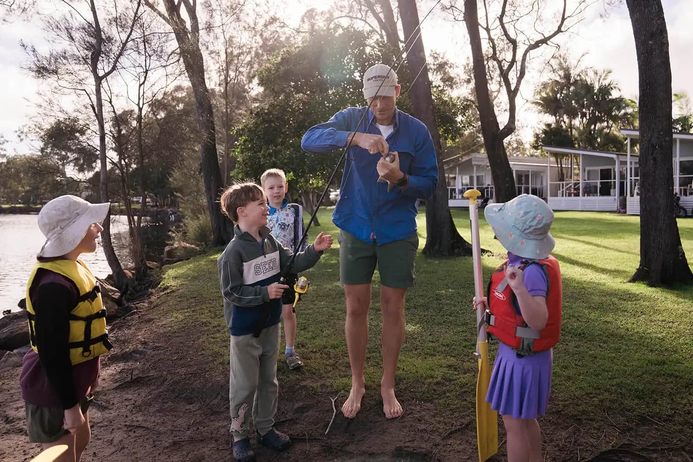 A dad and three children fishing from the grassy riverbank at Solitary Islands Resort, cabins and palms behind them in afternoon light