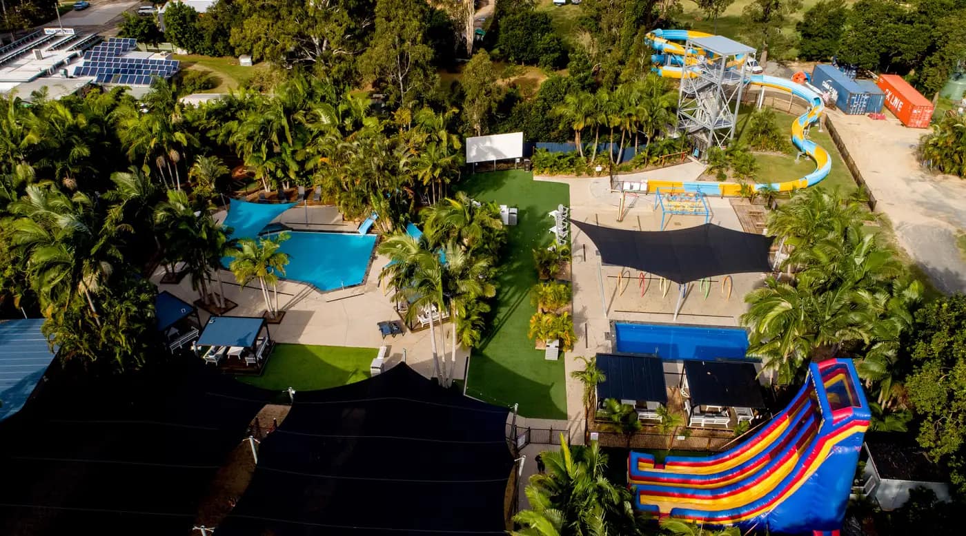 Aerial view of the resort's water play area: two swimming pools among palms, shade sails over the paths, and the tall waterslide towers with a striped inflatable slide beside them.