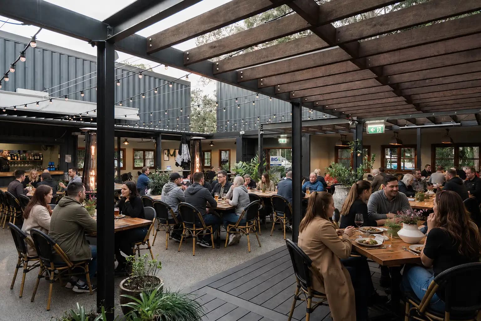 The 383's covered deck at full tilt: guests eating and drinking at long timber tables and black rattan chairs under a hardwood pergola strung with festoon lights, with the bar and dining room open behind.