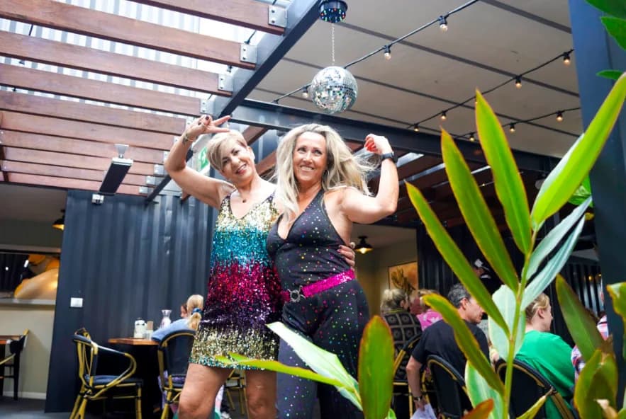 Two guests in sequinned outfits striking a pose under a mirror ball on music bingo night, with diners at tables behind them.