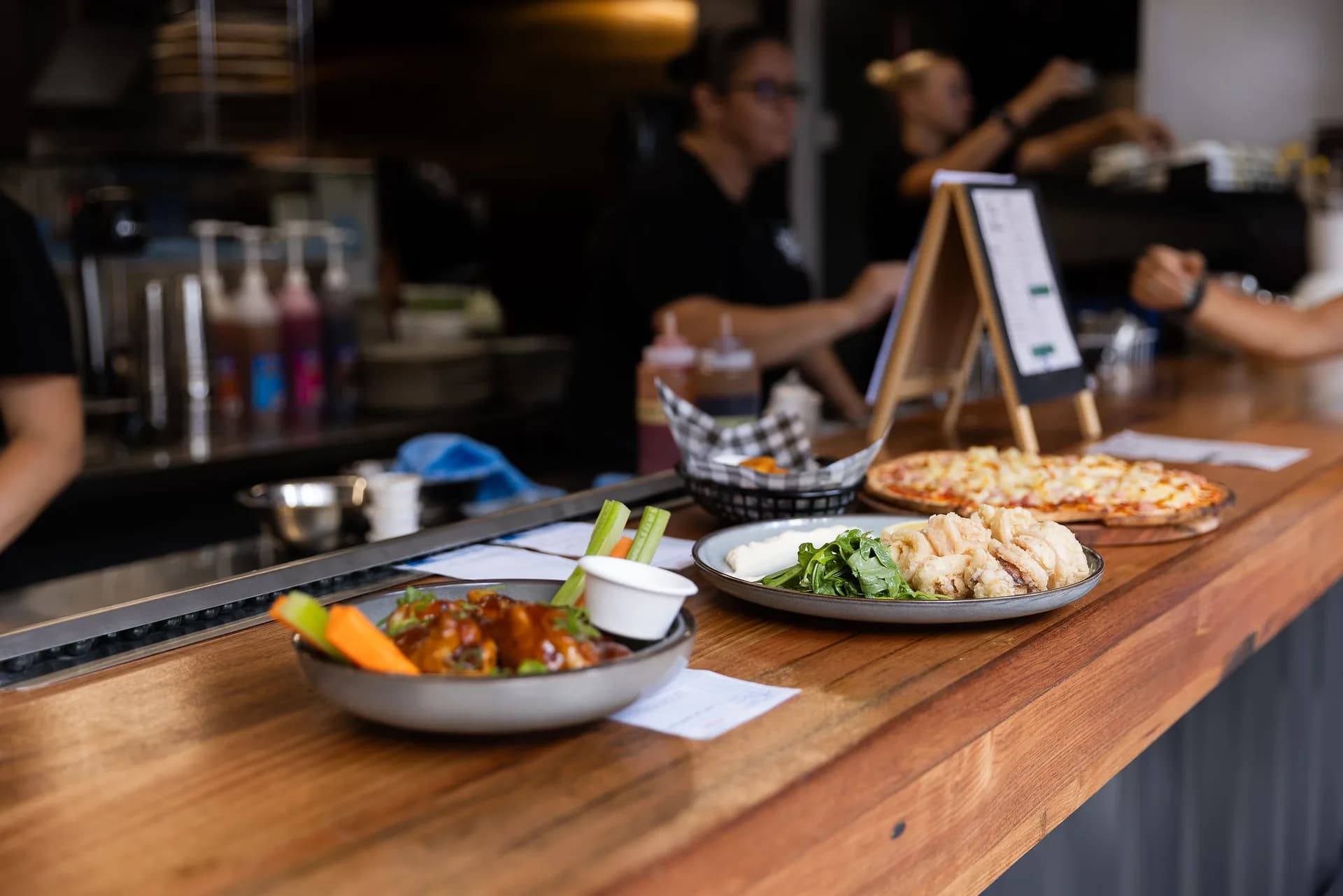 Plates lined up along the kitchen pass: saucy chicken wings with celery and carrot sticks, fried calamari with rocket and aioli, and a pizza waiting behind.