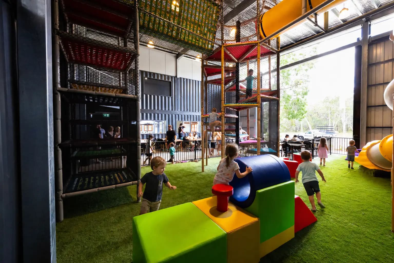 Inside The Play Pen: toddlers clambering over bright green, red and yellow soft-play blocks and a padded blue tunnel on artificial turf, with multi-storey netted climbing towers and a yellow tube slide behind them.