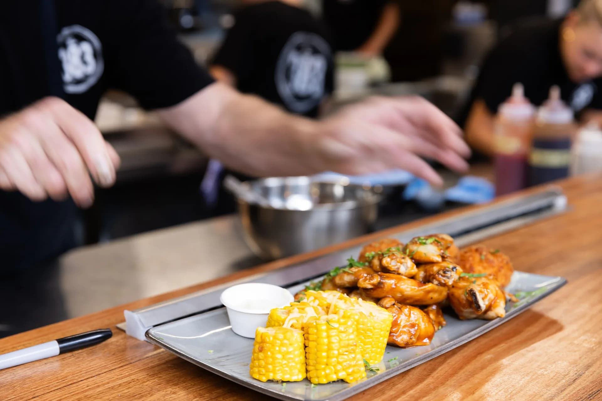 A share plate of glazed chicken wings with cheesy corn cobs and a pot of dipping sauce on the pass, kitchen staff working behind.