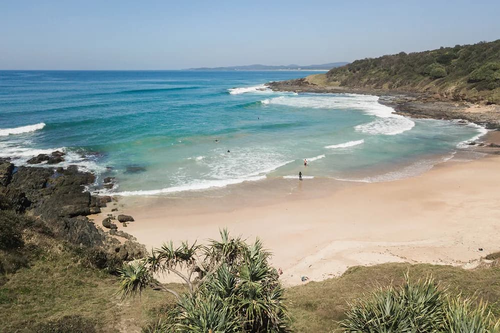 A sheltered sandy cove on the Yuraygir coastal walk seen from the headland: turquoise water breaking white over dark rocks, a few surfers in the shallows and pandanus palms in the foreground.