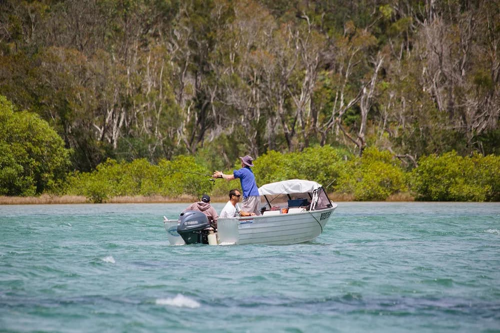 Three people fishing from a small aluminium tinnie on the Wooli Wooli River, one standing in a wide-brim hat to cast a line and two seated at the stern beside the outboard motor, with paperbark forest lining the far bank.