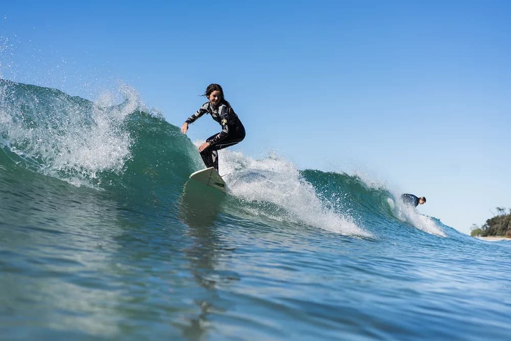 A young surfer in a wetsuit rides the face of a clean green wave off the Wooli coast on a bright day, with a second surfer paddling over the shoulder behind.