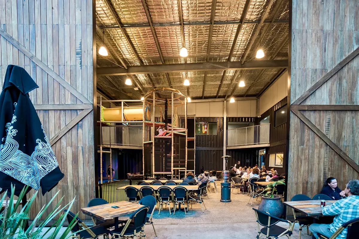 Tall timber barn doors opened wide onto the dining hall, with long share tables, diners and a multi-level rope climbing tower inside.