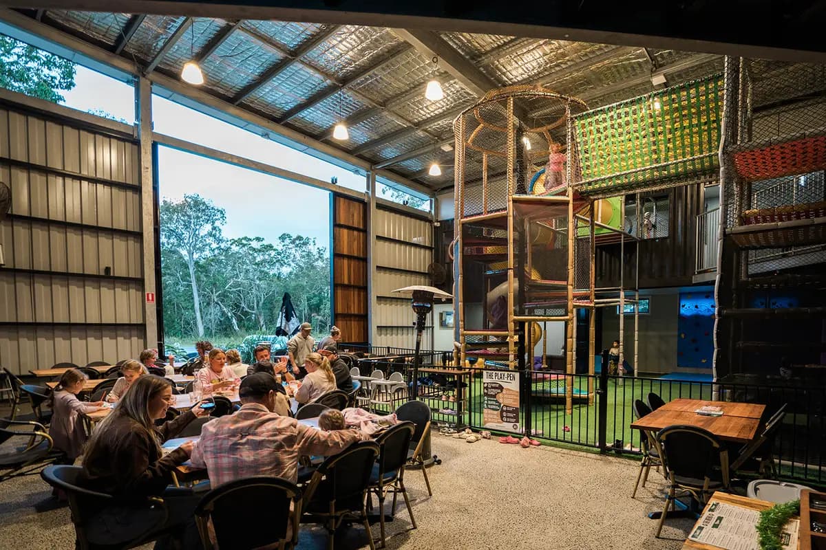 Families eating at tables in the open-sided dining shed at dusk, beside the fenced Play Pen climbing tower.