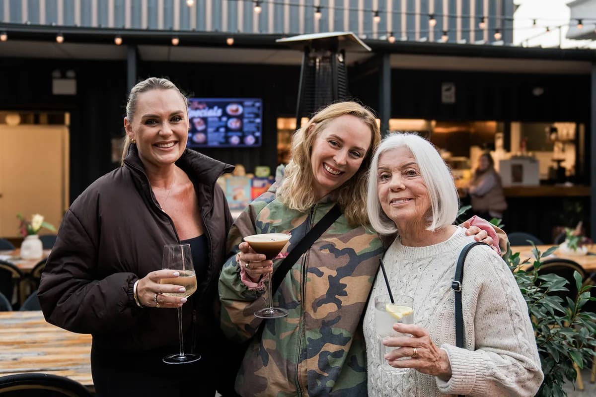 Three women smiling together with a wine, an espresso martini and a lemonade in hand at the outdoor bar, festoon lights strung overhead.