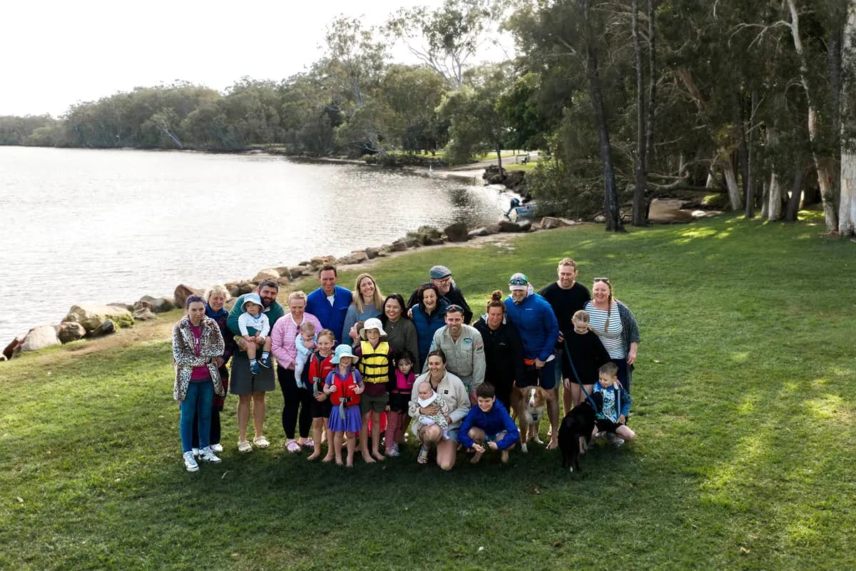 A big group of adults, kids and two dogs standing together on the grass beside the Wooli Wooli River, bush treeline on the far bank.