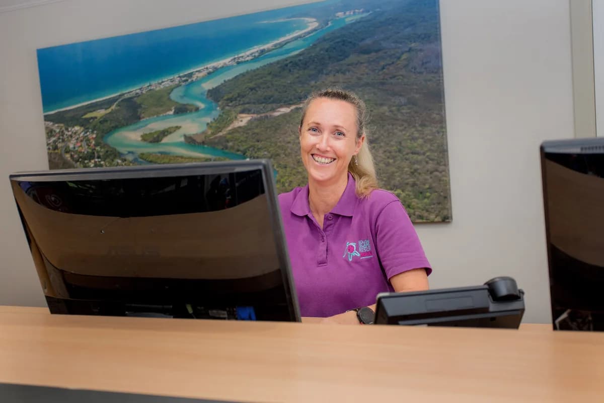 A resort team member in a purple polo smiling behind the reception desk, an aerial print of the Wooli River mouth on the wall behind her.