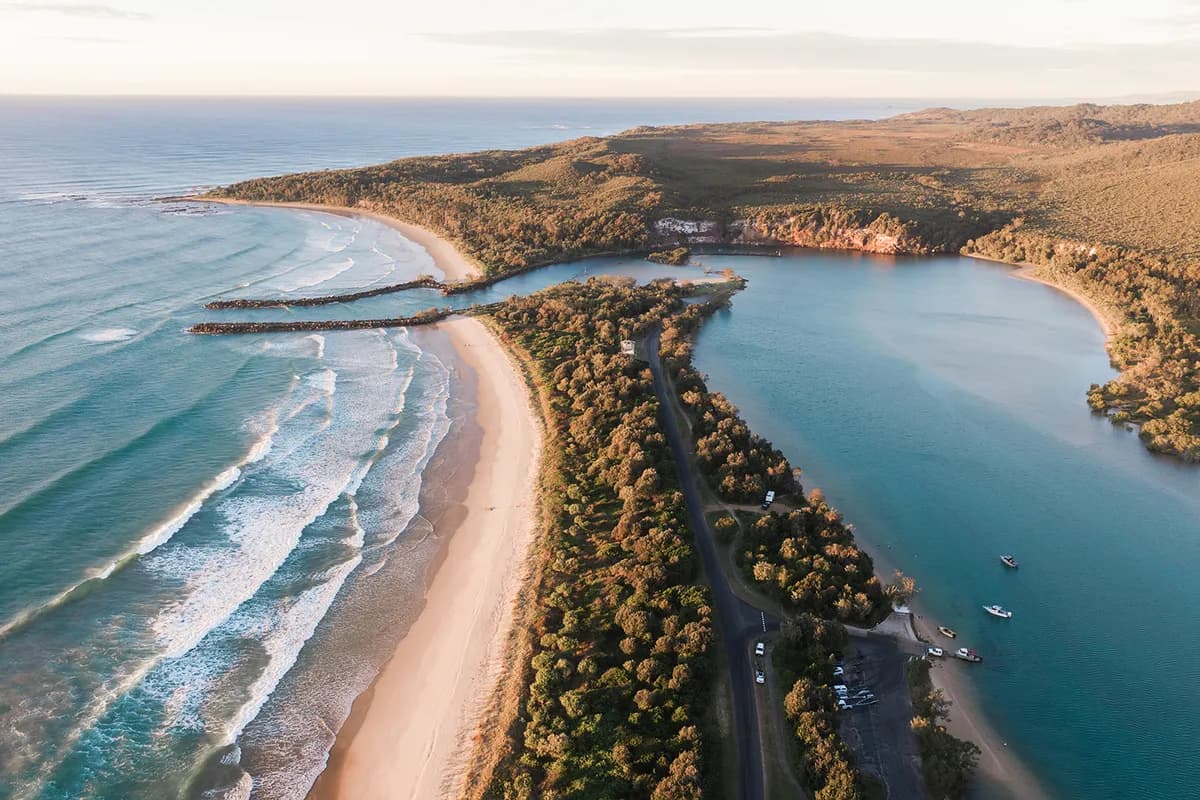 Sunset aerial of the Wooli River mouth meeting the ocean: surf rolling onto the beach, rock breakwalls and boats moored in the channel.