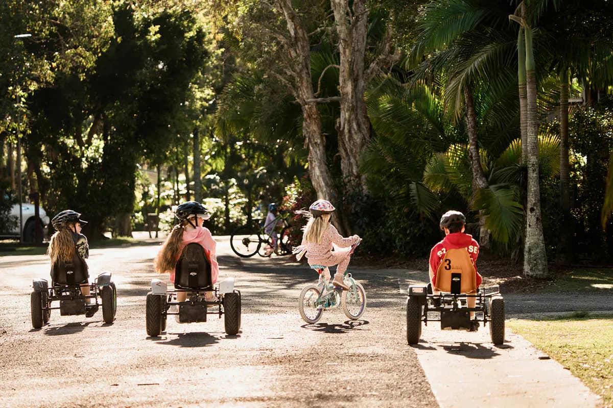 Three kids on pedal go-karts and one on a bike riding away down a shaded, palm-lined road through the park.