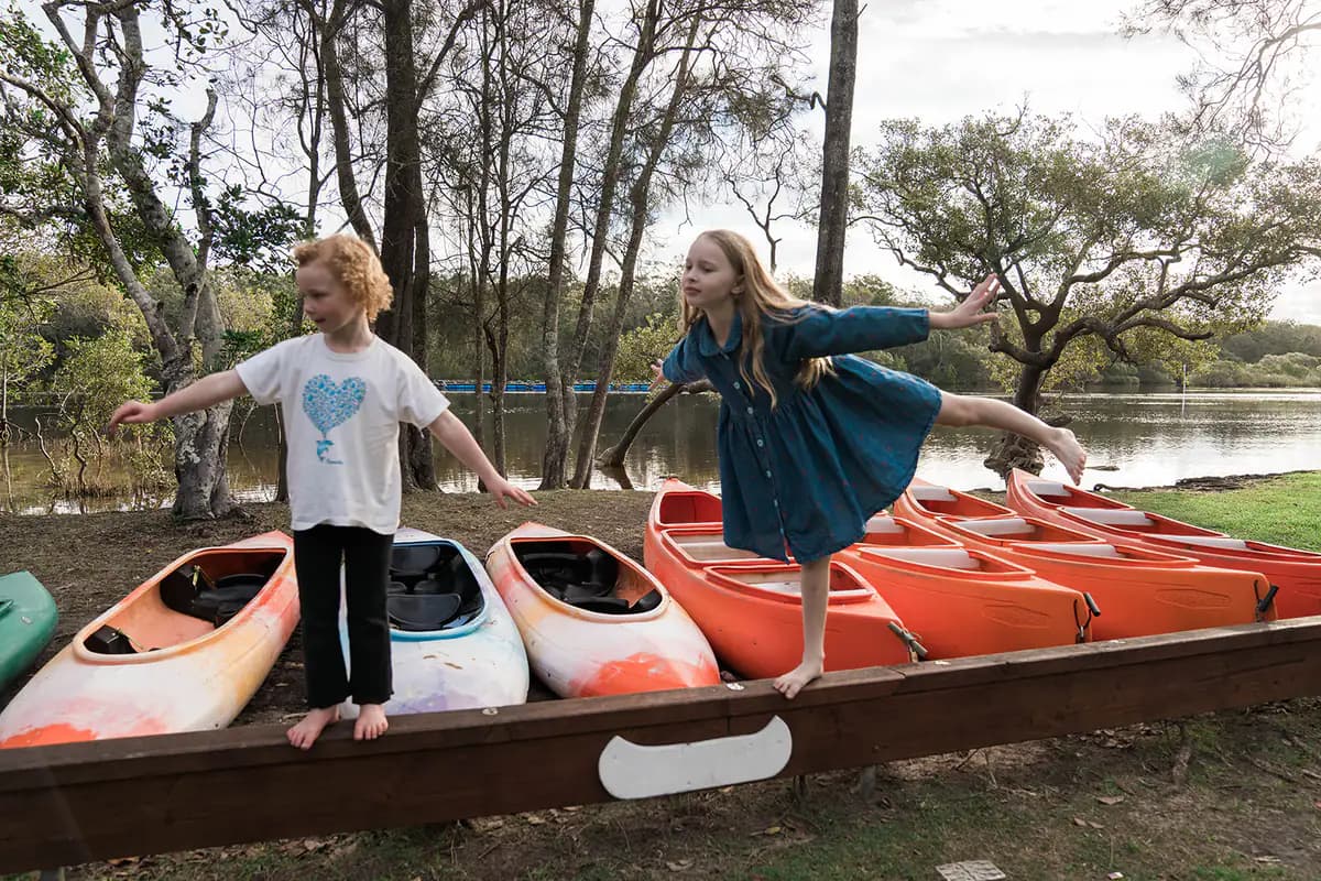 Two barefoot kids balancing along a timber beam in front of a row of orange kayaks and canoes by the river.