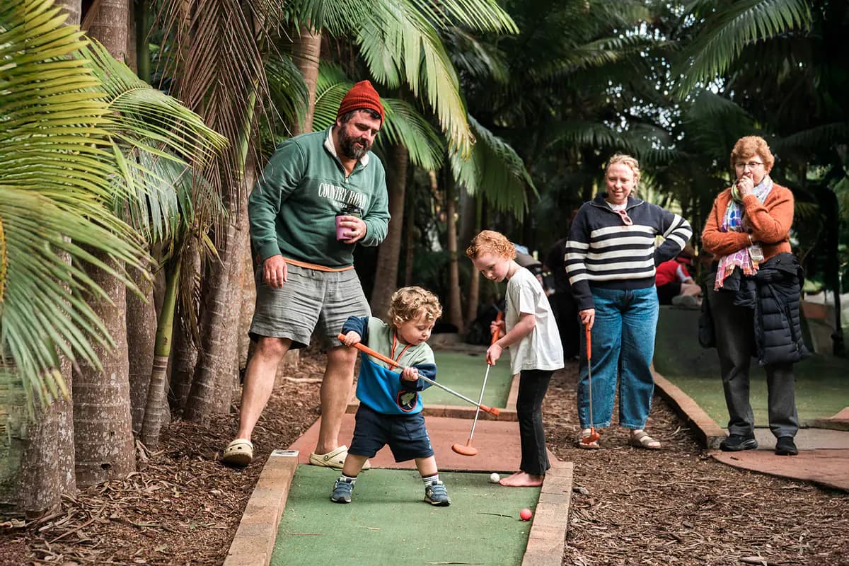 A toddler taking a putt on the mini golf green under tall palms while the rest of the family watches.