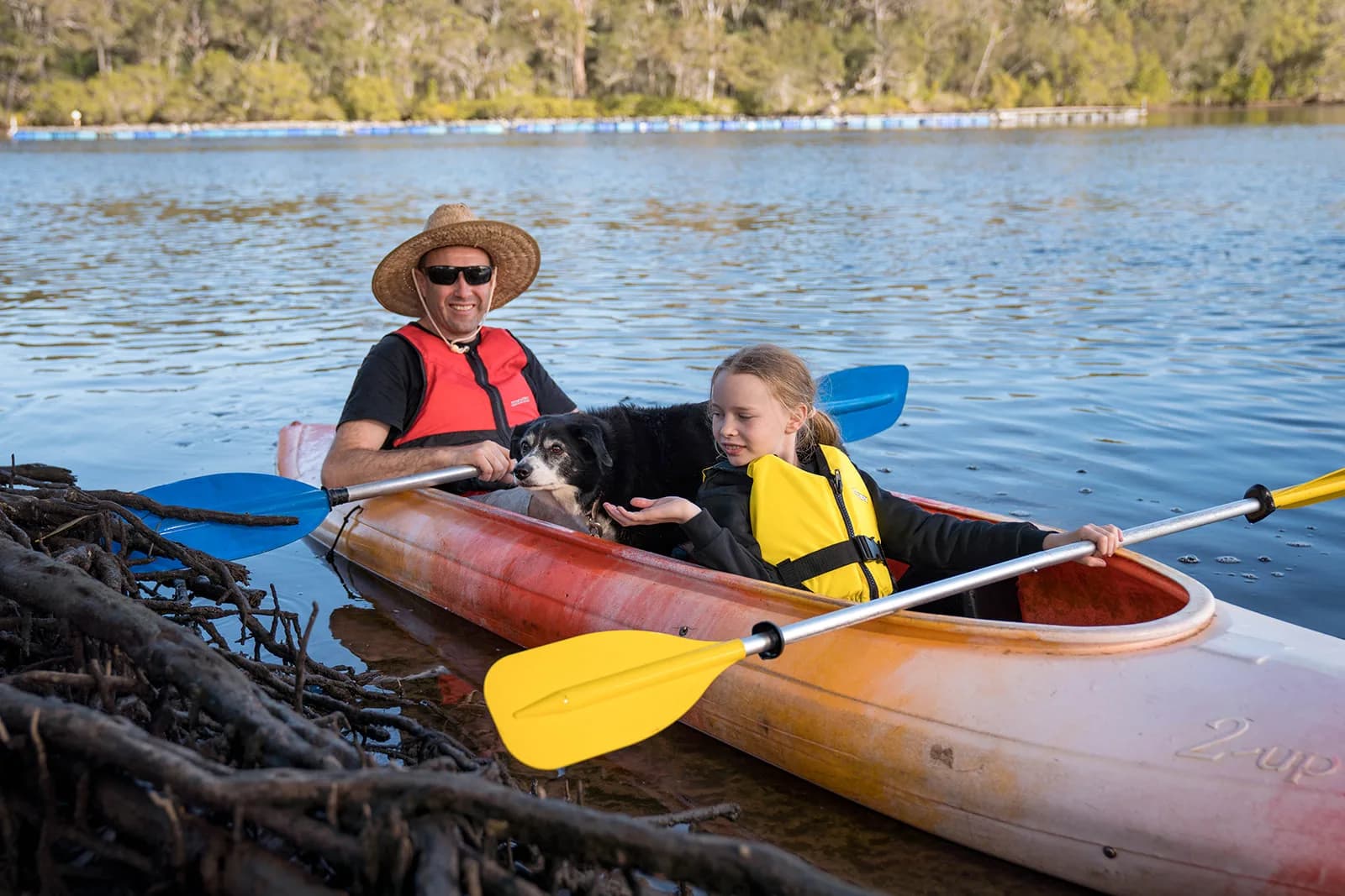 A man in a straw hat and sunglasses and a young girl in a yellow life jacket paddle a two-seater kayak on the Wooli Wooli River, a black and white dog sitting between them.