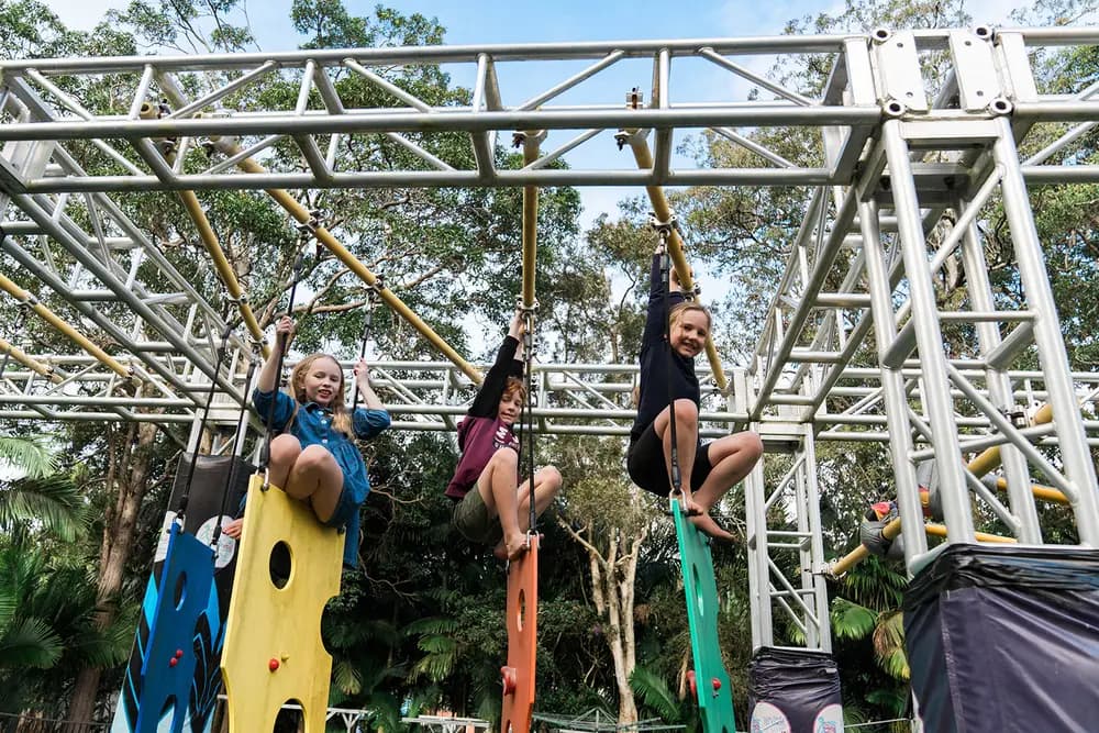 Three children hang from the overhead rings of the Wooli Warrior obstacle course, feet braced on coloured climbing panels below the steel truss frame, with tall gum trees behind.