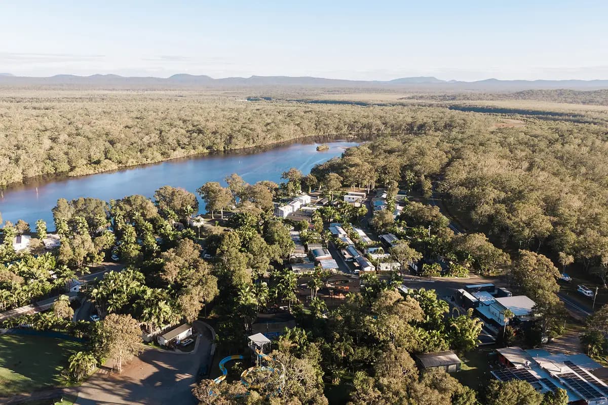 Aerial view over the resort: homes and caravan sites set among palms and gum trees on a bend of the Wooli River, the waterslide at the park's edge, and forest running to the ranges beyond.