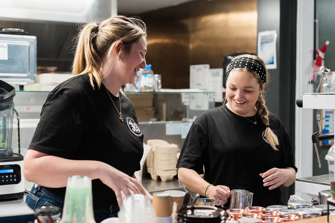 Two Solitary Islands Resort team members laughing together behind the counter at 383 Bar and Restaurant, one steaming milk at the coffee machine