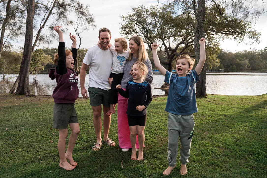 A family of six on the riverbank lawn at golden hour, the kids barefoot with their arms thrown in the air.