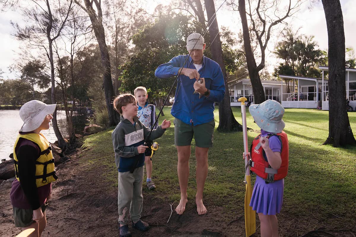 A dad unhooking a small fish on the riverbank while three kids in life jackets and sun hats watch with their rods.