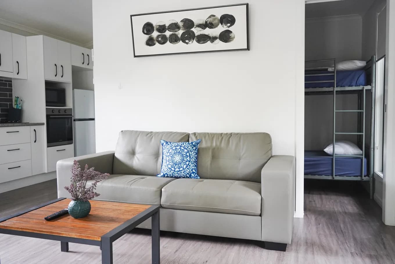 Lounge in the Two Bedroom Accessible Cabin, with a grey two-seater sofa and a timber coffee table, the kitchen to one side and the bunk room through the doorway beyond.