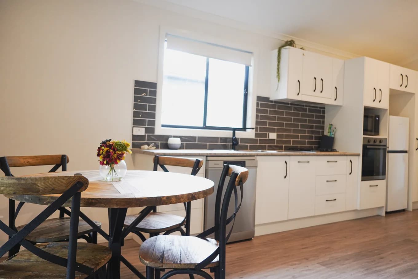 Kitchen and dining in the accessible cabin: white cabinetry with a brick-look splashback, a wall oven and dishwasher, beside a round timber table with metal chairs.