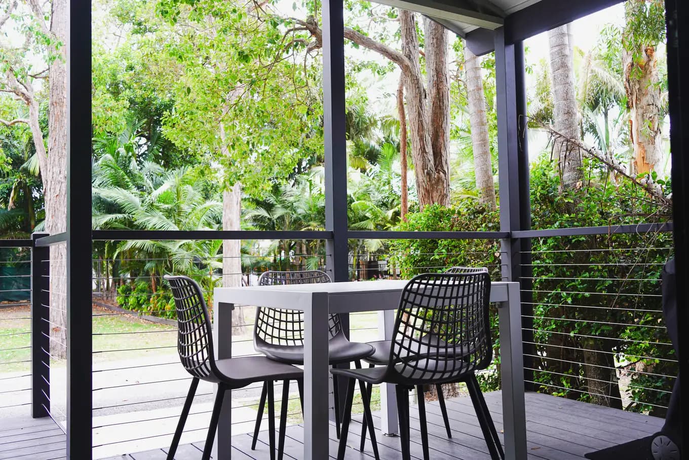 Deck at the accessible cabin, with a table and three black wire chairs behind a wire balustrade, screened by palms and subtropical greenery.