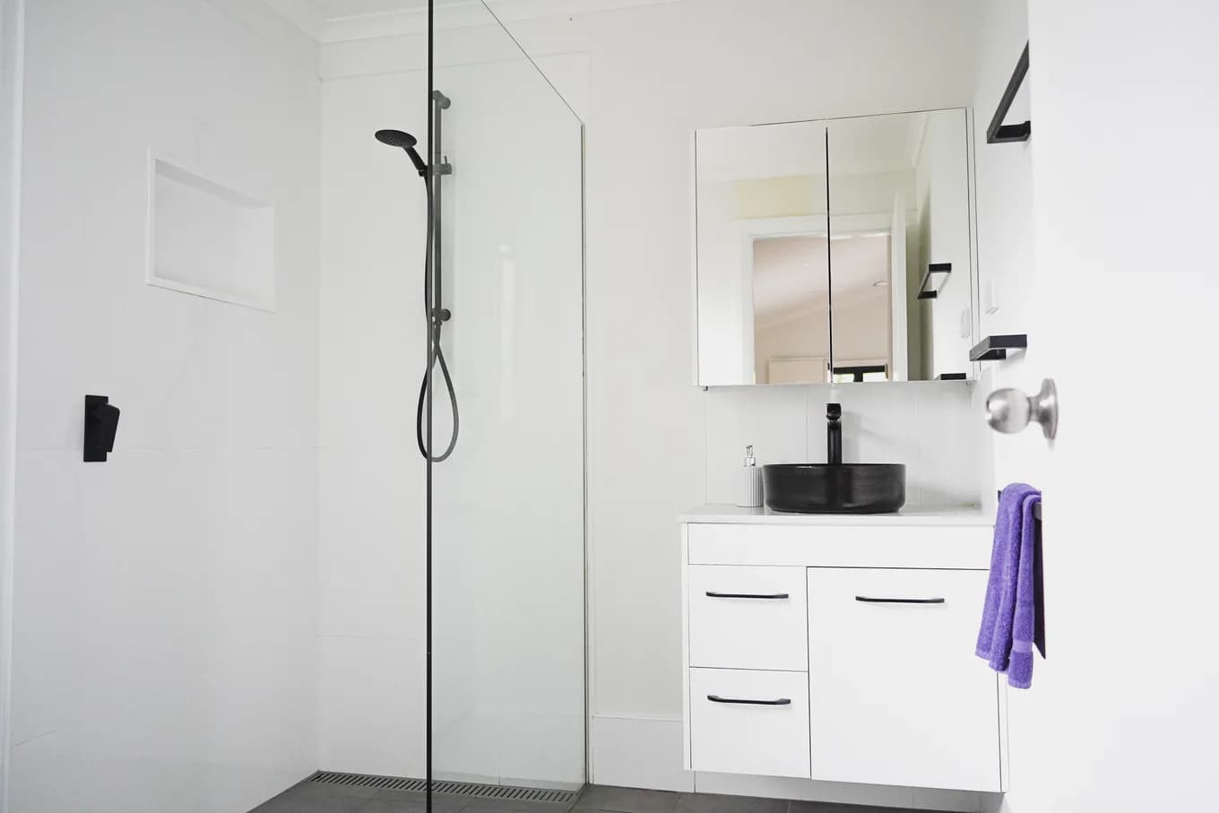Roll-in shower in the accessible cabin with no hob at the entry, alongside a white vanity with a black basin and a mirrored cabinet.