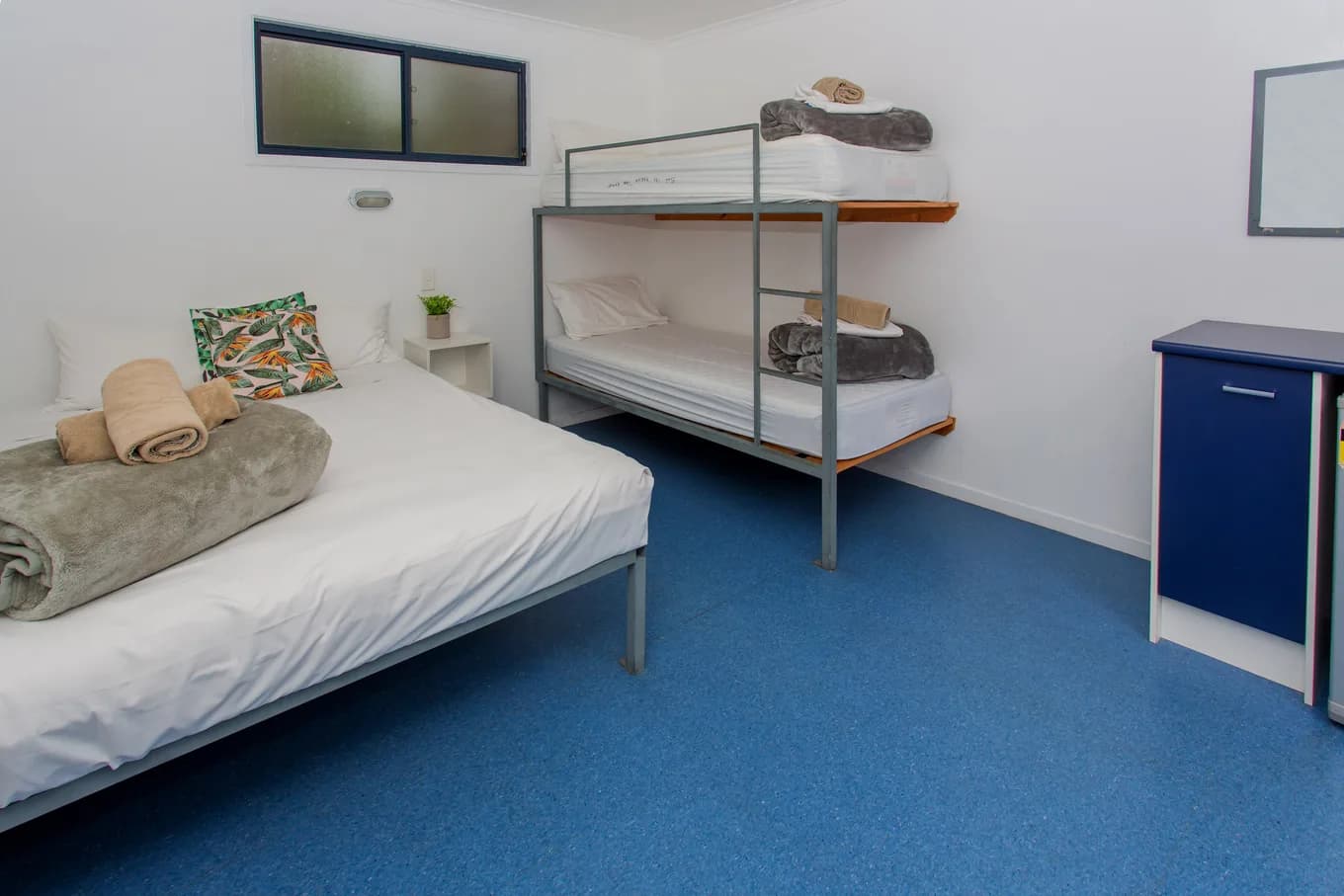 Beach Hut interior with a double bed made up with rolled towels and a patterned cushion, a metal bunk alongside, and a blue storage cabinet against the wall.
