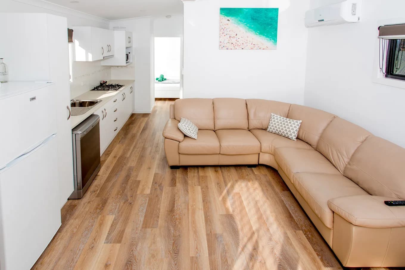 Living room in the Executive Two Bedroom Cabin, with a large tan leather corner lounge, the galley kitchen beyond and an aerial beach print on the wall.