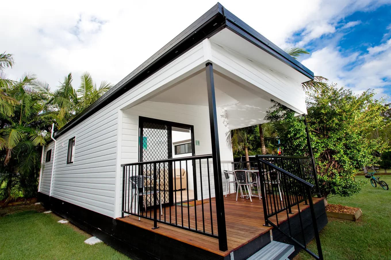 Exterior of the Executive Two Bedroom Cabin, white weatherboard with a covered timber deck, outdoor table and chairs, and palms behind.