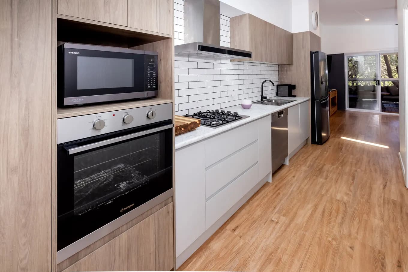 Galley kitchen in the villa: timber-look cabinetry, white subway-tile splashback, gas cooktop, wall oven and microwave, dishwasher and a black french-door fridge, opening through to the lounge.