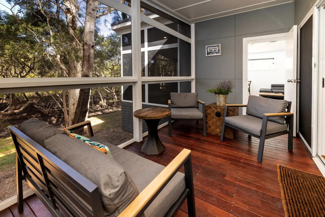 Screened deck off the villa, with a timber floor, a cushioned lounge and two armchairs around a low round table, looking out into the paperbarks.