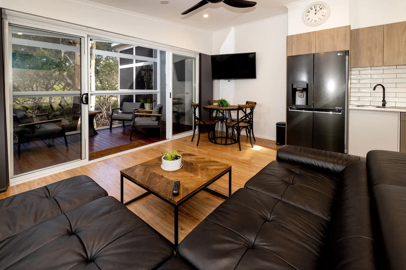 Open-plan living area in the villa: black leather lounges around a timber coffee table, a wall-mounted TV, the dining table and kitchen behind, and sliding doors out to the screened deck.
