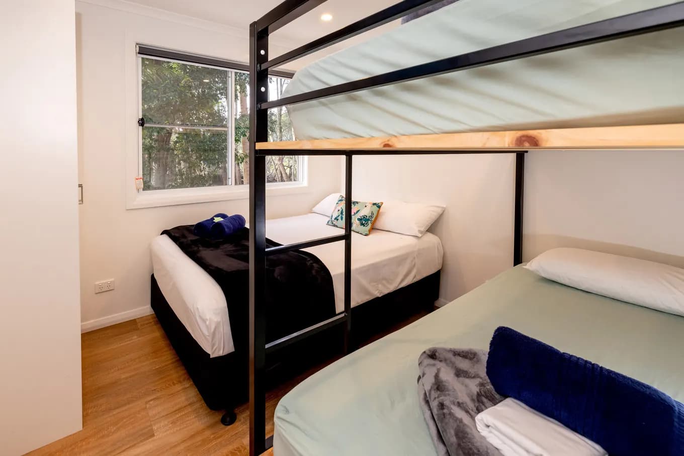 Bunk room in the villa, with a black steel bunk bed beside a single bed made up in white linen, and a window onto the trees.