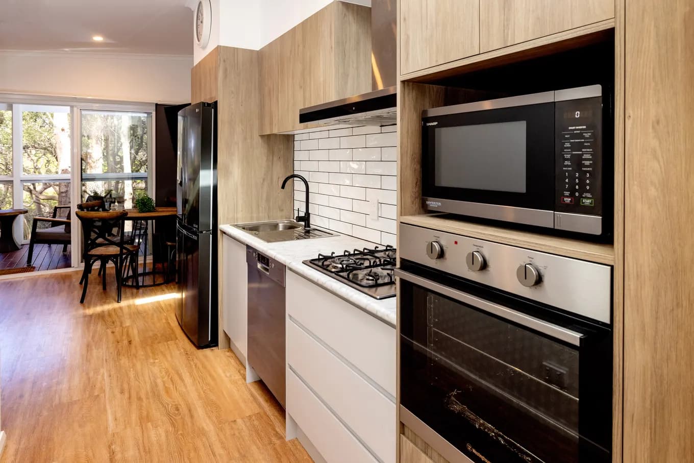 The villa kitchen looking the other way: wall oven, microwave, gas cooktop, dishwasher and fridge along a timber-floored galley, with the dining nook and windows beyond.