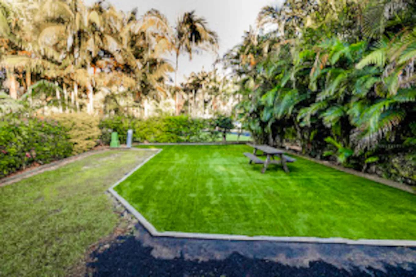 Wide rectangular powered turf site edged in concrete, with a picnic table at the far end and dense palms behind.