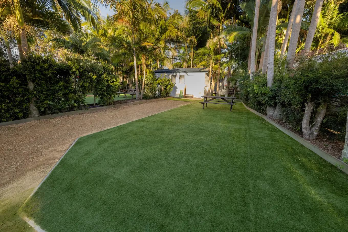 Powered turf site fringed by tall palms, with a gravel apron at the front and a picnic table at the far end.