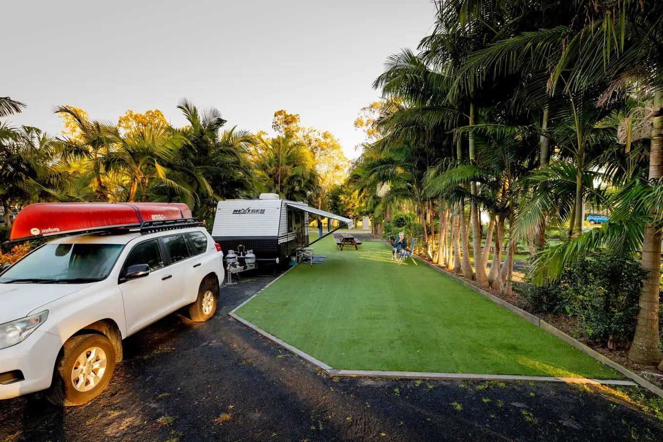 A four-wheel drive with a red canoe on its roof parked alongside a caravan on a powered turf site, framed by tall palms.