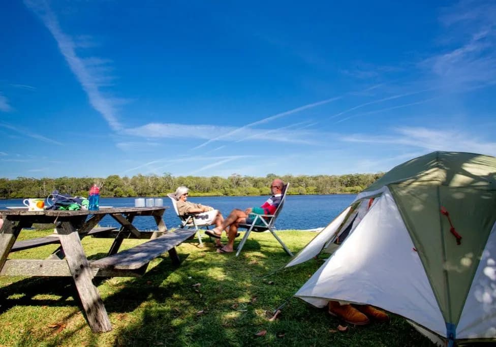 Two campers relaxing in folding chairs beside a picnic table and a pitched tent on a premium river site, with the river stretching out behind them.