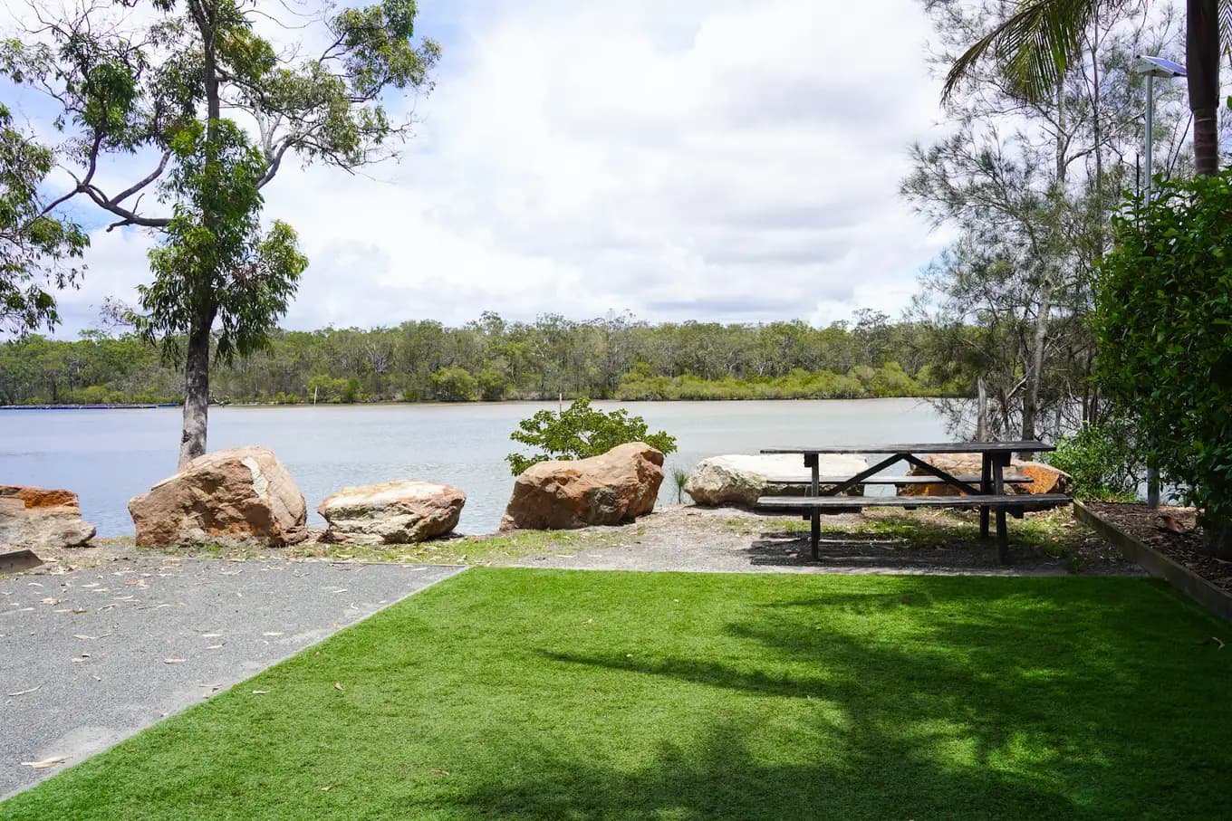 Picnic table on the rocky river bank at a riverfront tent site, with the turf pad in the foreground and the water beyond.