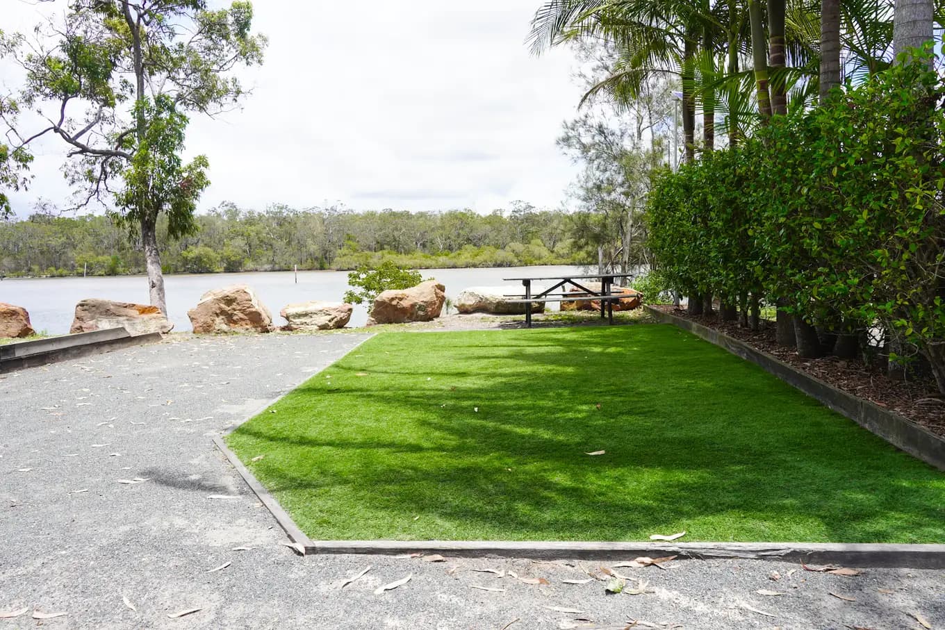 Turf pad at a riverfront site beside a gravel driveway, with room for a small campervan and the river through the trees.