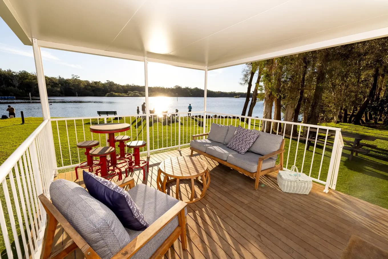 Covered riverfront deck at the Riverview Three Bedroom Villa, with a timber bar table and red stools, a cushioned sofa, and the lawn running down to the water at sunset.