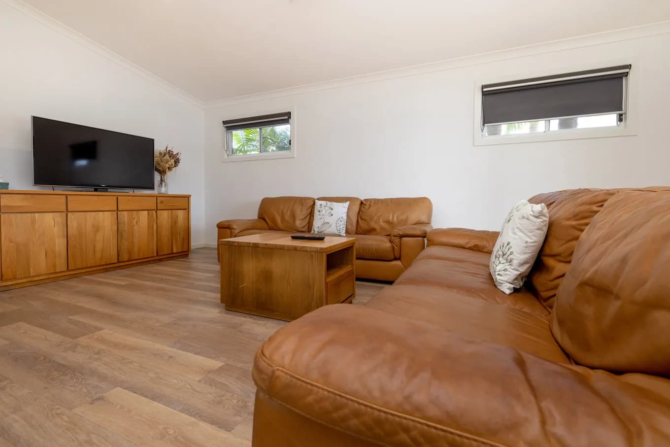 Living room in the three bedroom villa, with a large tan leather corner lounge, a wall-mounted TV and a timber sideboard on timber-look floors.
