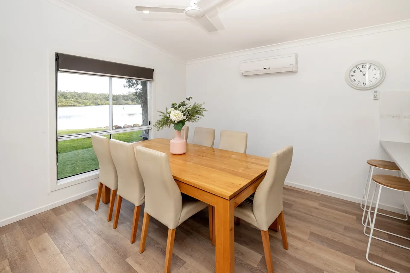 Dining area in the three bedroom villa: a six-seat timber table with cream upholstered chairs beside a wide window looking out over the lawn to the river.