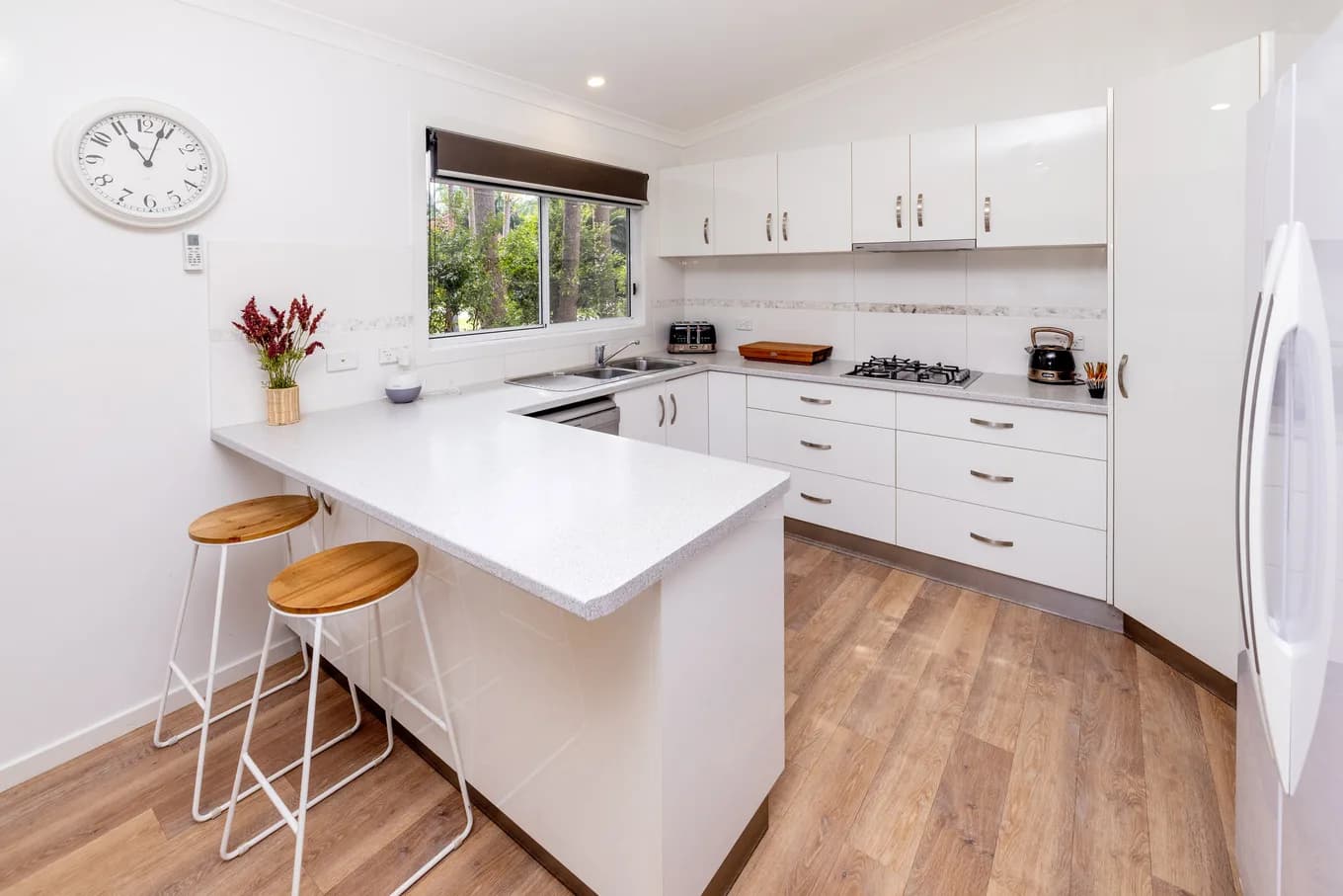 U-shaped white kitchen in the three bedroom villa, with a stone breakfast bar and two timber stools, a gas cooktop, and a full-height fridge.